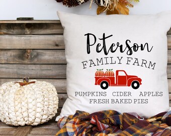 May include: A white throw pillow with the text "Peterson Family Farm" and a red truck carrying pumpkins. The text below the truck reads "Pumpkins Cider Apples Fresh Baked Pies". The pillow is on a wooden crate with a white pumpkin and a plaid blanket.