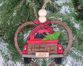 May include: A wooden heart-shaped Christmas ornament with a red truck carrying a Christmas tree. The truck has a red bow on the hood and the text "Heather & Tom 2023" on the back.