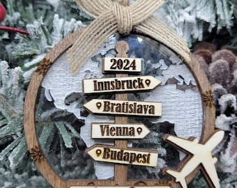 May include: A wooden Christmas ornament with a world map design and a plane silhouette. It features a signpost with the names of cities: Innsbruck, Bratislava, Vienna, and Budapest. The ornament also includes the year 2024 and the names "Cy & Dave".