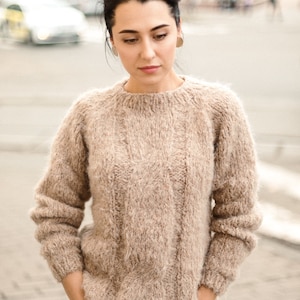 May include: A woman wearing a light brown fuzzy sweater with a cable knit pattern and blue jeans. She is standing on a brick walkway.
