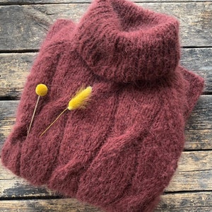 Puede incluir: Un suéter de cuello alto color marrón rojizo hecho de un hilo suave y peludo. El suéter está doblado y colocado sobre una superficie de madera.
