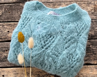 Può includere: Un maglione di maglia blu chiaro e soffice con scollo tondo e design traforato. Il maglione è piegato su una superficie di legno rustica, con fiori secchi in primo piano. L'etichetta recita "KOSA".