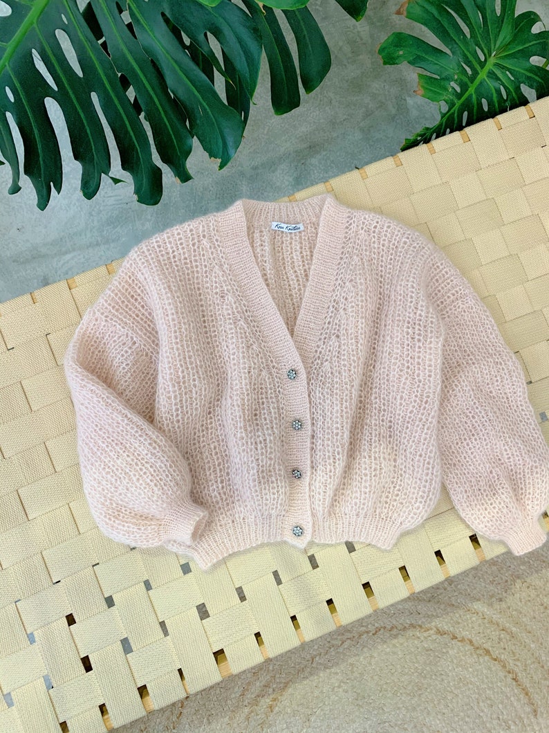 K&ouml;nnte beinhalten: Ein hellrosa gestrickter Cardigan mit V-Ausschnitt und Kn&ouml;pfen. Der Cardigan hat eine leicht verk&uuml;rzte L&auml;nge und eine lockere Passform.