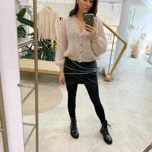 K&ouml;nnte beinhalten: Eine Frau tr&auml;gt einen hellrosa, flauschigen Cardigan, einen schwarzen Lederrock, schwarze Strumpfhosen und schwarze Combat Boots. Sie h&auml;lt ein Handy in ihrer rechten Hand und macht ein Selfie.