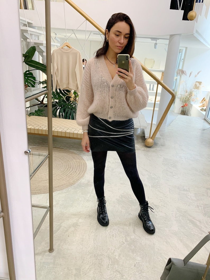 K&ouml;nnte beinhalten: Eine Frau tr&auml;gt einen hellrosa, flauschigen Cardigan, einen schwarzen Lederrock, schwarze Strumpfhosen und schwarze Combat Boots. Sie h&auml;lt ein Handy in ihrer rechten Hand und macht ein Selfie.