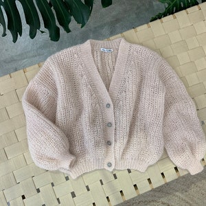 K&ouml;nnte beinhalten: Ein hellrosa gestrickter Cardigan mit V-Ausschnitt und Kn&ouml;pfen. Der Cardigan hat eine lockere Passform und eine leicht verk&uuml;rzte L&auml;nge. Die Kn&ouml;pfe sind dekorativ und haben eine perlmuttartige Oberfl&auml;che.