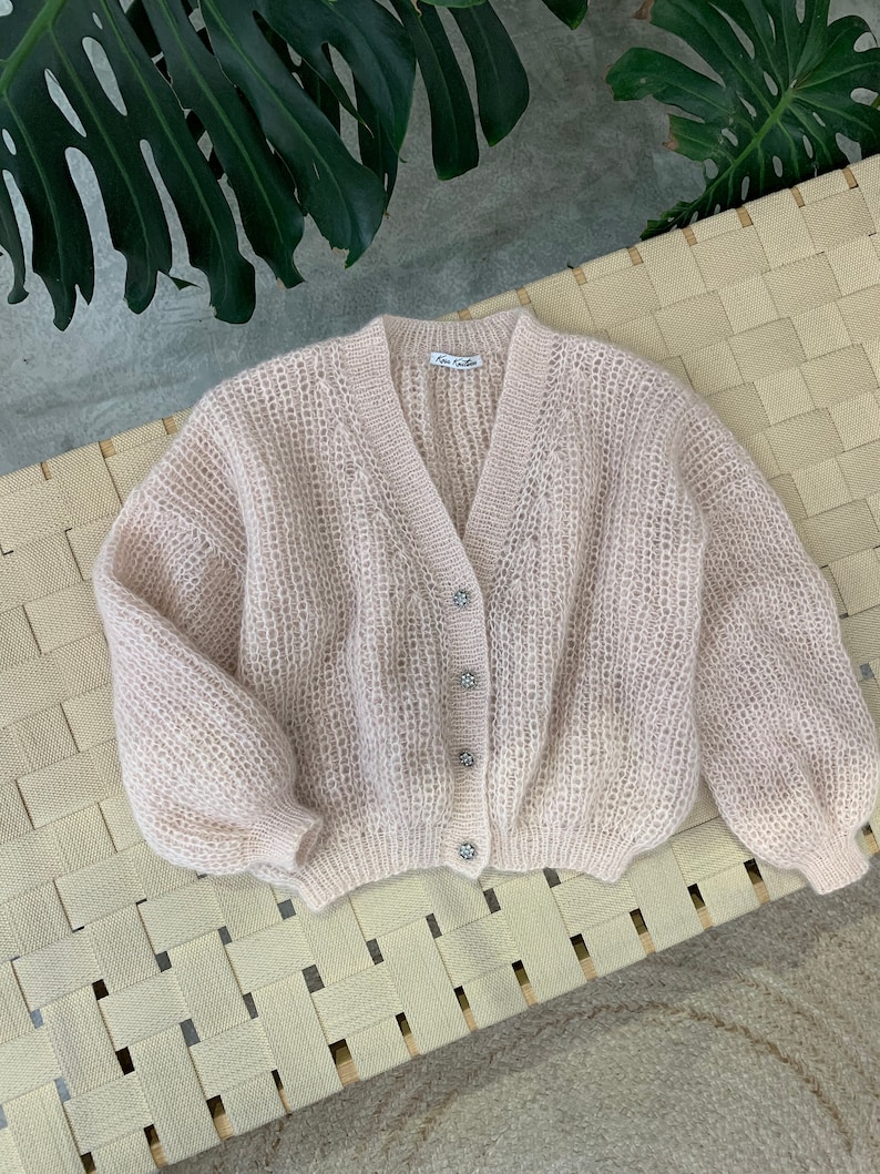 K&ouml;nnte beinhalten: Ein hellrosa gestrickter Cardigan mit V-Ausschnitt und Kn&ouml;pfen. Der Cardigan hat eine lockere Passform und eine leicht verk&uuml;rzte L&auml;nge. Die Kn&ouml;pfe sind dekorativ und haben eine perlmuttartige Oberfl&auml;che.