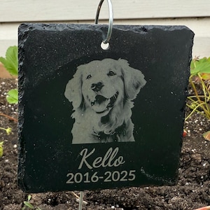 May include: A black slate pet memorial with a silver hanging loop. The memorial features an engraved image of a Golden Retriever dog with the name "Kello" and the years "2016-2025" in white lettering. The memorial is square-shaped.