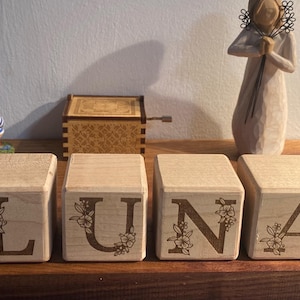 Peut inclure: Quatre blocs de bois avec les lettres L, U, N, A épellées. Chaque bloc a un motif floral gravé sur le devant.