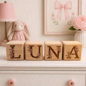 May include: Four light-coloured wooden blocks arranged in a row, each featuring a dark, engraved letter that spells out LUNA. Each letter is decorated with a detailed floral vine pattern etched into the wood surface of the square blocks.