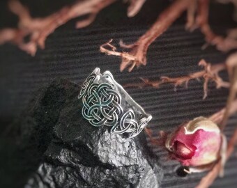 Puede incluir: Un anillo de plata con un diseño de nudo celta, descansando sobre una roca negra. El anillo está rodeado por una rama seca con un pequeño capullo de flor rosa.