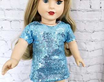 May include: A doll with blonde hair and blue eyes wearing a short-sleeved, blue sequin dress and black glitter boots. The doll has red lipstick and is posed against a white brick wall.