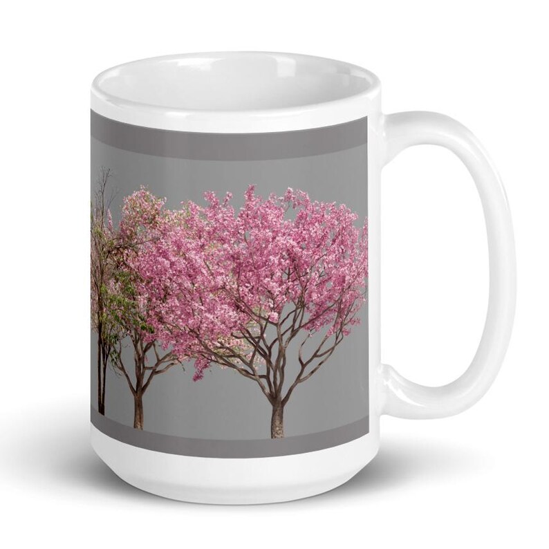 May include: White ceramic mug with a gray background and a design of three pink flowering trees. The trees are in a row and have green leaves.