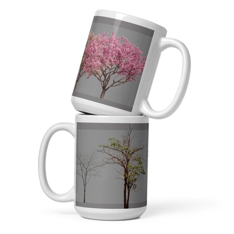 May include: Two white ceramic mugs with a gray background. The top mug features a pink flowering tree. The bottom mug features a tree with green leaves and a bare tree.