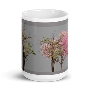 May include: White ceramic mug with a gray background and a design of three trees with pink and green leaves. The trees are arranged in a row, with the tree on the left being bare, the middle tree having pink leaves, and the right tree having green leaves.