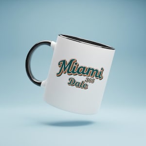 Puede incluir: Taza de cerámica blanca con asa y borde negros. La taza presenta las palabras "Miami 305 Dale" en escritura turquesa y naranja. La taza está sobre un fondo azul claro.