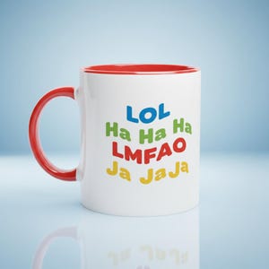 May include: White ceramic mug with a red handle and interior. The mug features the text "LOL Ha Ha Ha LMFAO Ja Ja Ja" in blue, green, red, and yellow. The mug is on a light blue surface.