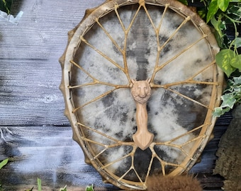 May include: A brown leather drum with a wooden wolf head handle. The drum has a white background and a intricate design of brown leather strips.