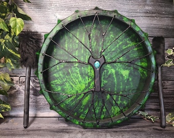 May include: A round frame drum with a vibrant green and black mottled surface, featuring a central black handle adorned with a small turquoise stone. The drum is laced with black cord in a radial pattern and includes two black fur-tipped mallets.
