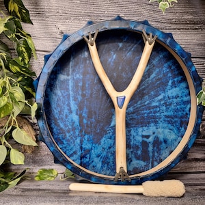 May include: A blue hand drum with a wooden handle and a blue gemstone. The drum is surrounded by a wooden frame with a decorative edge. A wooden stick and a white brush are in the foreground.
