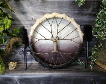 May include: A round, hand-crafted drum with a tree of life design painted on the front. The drum is made of wood and leather and has a dark brown and white color scheme. It comes with two black drum beaters.