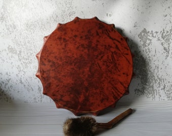 Può includere: Un tamburo in pelle marrone con un telaio in legno e un battitore rivestito in pelliccia marrone. Il tamburo presenta un motivo decorativo sulla pelle.