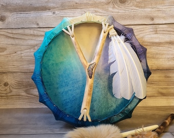 May include: A blue, green, and purple hand drum with a wooden Y-shaped handle and white feathers. The drum is decorated with a gradient of colors and has a leather surface.