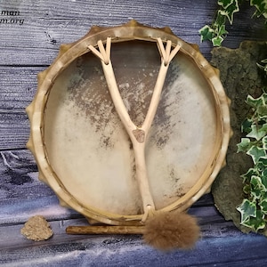 Könnte beinhalten: Eine braune Ledertrommel mit einem Holzrahmen und einem Y-förmigen Griff. Die Trommel ist mit einem kleinen Stück Holz in der Mitte des Griffs verziert. Die Trommel wird von einem Holzstock und einem braunen, mit Fell überzogenen Schläger begleitet.