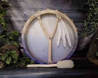 May include: A purple and white hand drum with a wooden Y-shaped handle and white feathers. The drum is accompanied by a wooden beater with a white brush.