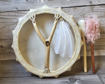 May include: A white frame drum with a wooden Y-shaped beater and white feathers. The drum is on a wooden surface with two drum beaters, one with a pink fluffy head and the other with a white fluffy head.