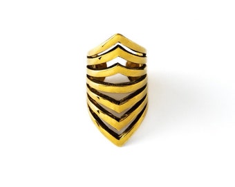 Puede incluir: Un anillo dorado con un diseño único. El anillo presenta múltiples bandas en forma de chevrón apiladas, creando un efecto de capas. El anillo está sobre un fondo blanco, resaltando sus intrincados detalles y brillo metálico. Este anillo llamativo es un accesorio audaz.