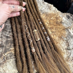 May include: Brown dreadlocks with decorative wraps and a gold-colored hair cuff. The dreadlocks are arranged in a row, with some wrapped in white, pink, and silver thread. The hair cuff is a gold color and has a textured design.