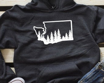 Può includere: Una felpa con cappuccio grigio scuro con un contorno bianco dello stato di Washington con alberi al suo interno. La felpa è appoggiata su una superficie di legno con un paio di scarpe da ginnastica bianche e un paio di jeans blu.