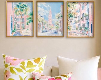 May include: A set of three vertical wall art prints in gold frames, each featuring a watercolor-style painting of a city street scene with pink buildings, palm trees, and a tall church steeple under a bright blue sky with white clouds.