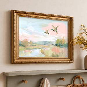 Könnte beinhalten: Gerahmtes Landschaftsgemälde mit zwei Enten im Flug über einem Sumpf. Das Kunstwerk verwendet weiche Pinselstriche und Pastellfarben, darunter Rosa, Blau und Grün, um eine ruhige Szene darzustellen. Der goldene Rahmen verleiht einen klassischen Touch.