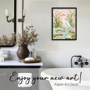May include: A framed painting of a white heron standing in a pond with lily pads and flowers. The artwork is displayed in a bathroom setting, with a mirror, vase, and bathtub visible. Text on the image reads "Enjoy your new art!" and "Aspen Art Decor."