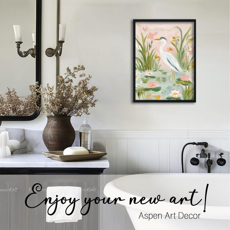 May include: A framed painting of a white heron standing in a pond with lily pads and flowers. The artwork is displayed in a bathroom setting, with a mirror, vase, and bathtub visible. Text on the image reads "Enjoy your new art!" and "Aspen Art Decor."