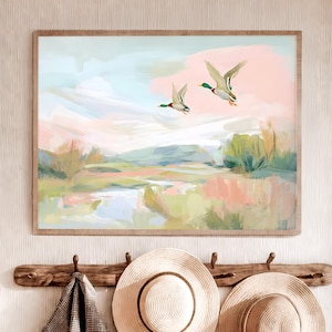 Op de afbeelding: Een ingelijst landschapsschilderij met een serene scène van een moeras en twee vliegende eenden. Het kunstwerk heeft zachte penseelstreken in pastelkleuren, waaronder roze, groen en blauw. Het schilderij wordt tentoongesteld aan een muur met een houten kapstok.