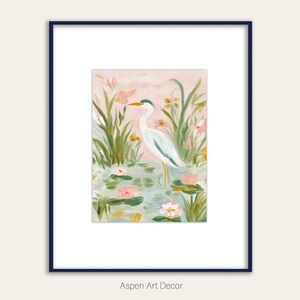May include: A framed art print featuring a serene watercolor painting of a white heron standing in a pond with lily pads and pink flowers. The artwork is set against a soft pink background, framed in a navy blue frame. The text "Aspen Art Decor" is at the bottom.