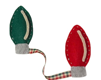 May include: Two felt Christmas light ornaments. One is green, the other is red, both with a white accent and gray base. They are connected by a plaid ribbon. The ornaments are designed to look like vintage Christmas lights.