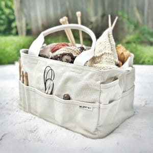 May include: A beige canvas tote bag filled with knitting supplies. The bag has multiple exterior pockets and a white handle. Inside are yarn balls, knitting needles, and a partially knitted item. The text "THE ARTIST NEST" is visible on a small tag.