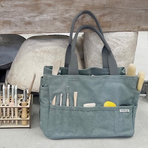Op de afbeelding: Een teal canvas tote bag met meerdere vakken, met diverse pottenbakkersgereedschappen. De tas heeft grijze handvatten en een klein wit label met de tekst "THE ARTIST NEST". Een houten gereedschapsorganizer en grote aardewerkstukken staan op de achtergrond.