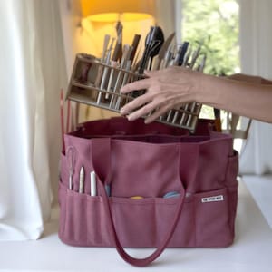 May include: A burgundy tote bag with multiple pockets and handles, holding a wooden organizer filled with art supplies. The bag has the text "THE ARTIST NEST" on the side. Various art tools are visible in the pockets.