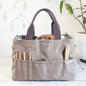 May include: A taupe canvas craft tote bag featuring two sturdy handles, multiple exterior pockets, and a small white label reading THE ARTIST NEST. The pockets contain several wooden crochet hooks and knitting needles, with a wooden hoop visible inside.