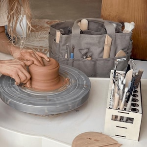 May include: A potter's wheel with a clay pot in progress, alongside a gray canvas tote bag filled with pottery tools. A wooden tool holder with various brushes and tools is also visible. The Artist's Nest is printed on the bag.