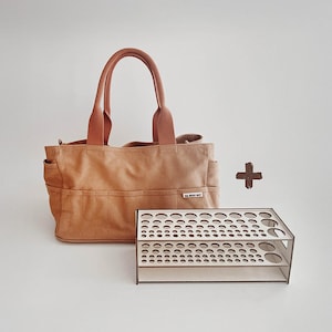 Pode incluir: Uma sacola de lona bege com alças marrons e vários bolsos externos. Ao lado, um organizador de madeira com recortes circulares de vários tamanhos. A sacola tem uma pequena etiqueta branca que diz "THE ARTIST NEST".