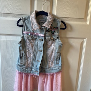 May include: A denim vest with a pink lace trim and a butterfly appliqué. The vest is layered over a pink tulle skirt with sequins.