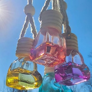 May include: Four heart-shaped glass car air fresheners in yellow, pink, purple, and teal, suspended by a rope. Each bottle has a wooden cap and is filled with a colorful liquid. The background is a clear blue sky.