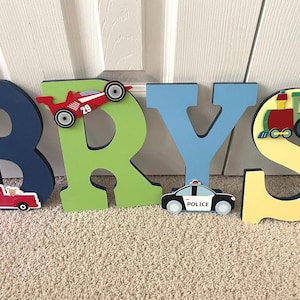 Puede incluir: Letras de madera que deletrean el nombre "BRYS" en varios colores. Cada letra est&aacute; decorada con un veh&iacute;culo diferente: un coche de carreras rojo, una camioneta roja, un coche de polic&iacute;a y un tren.