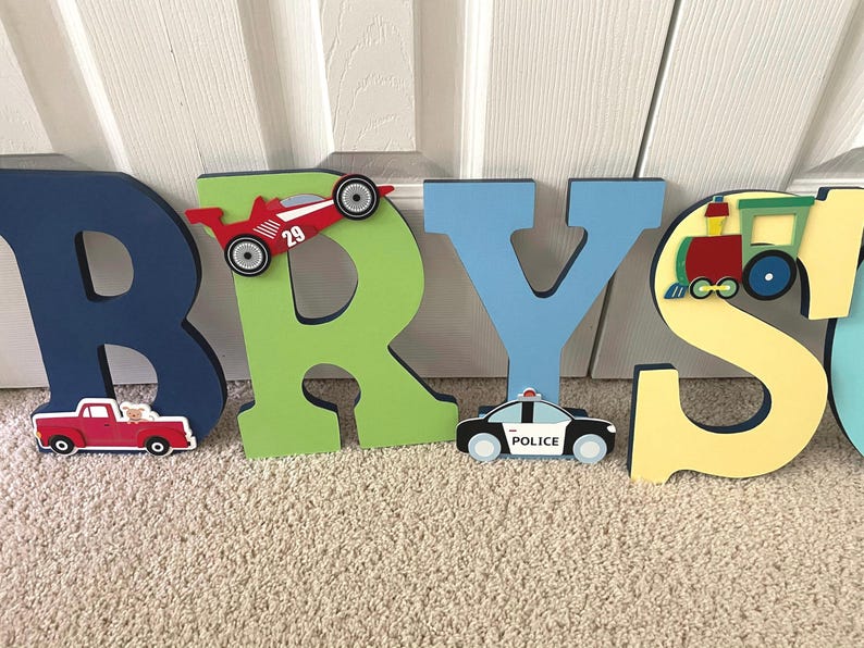 Puede incluir: Letras de madera que deletrean el nombre "BRYS" en varios colores. Cada letra est&aacute; decorada con un veh&iacute;culo diferente: un coche de carreras rojo, una camioneta roja, un coche de polic&iacute;a y un tren.